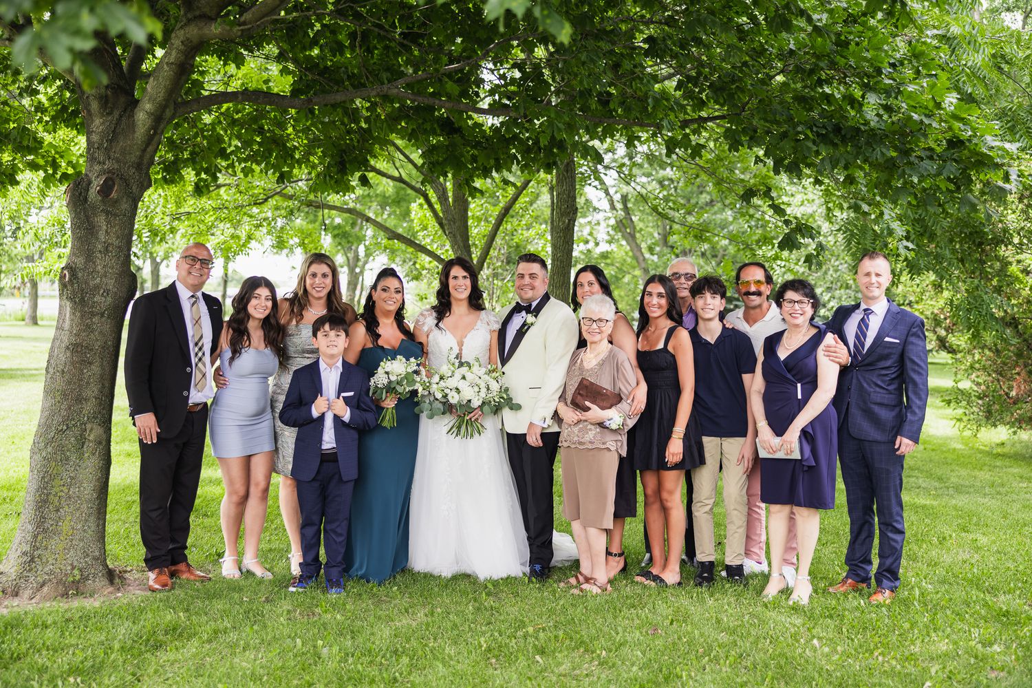 Wedding party group portrait under shady tree, bride and groom centered with family members.