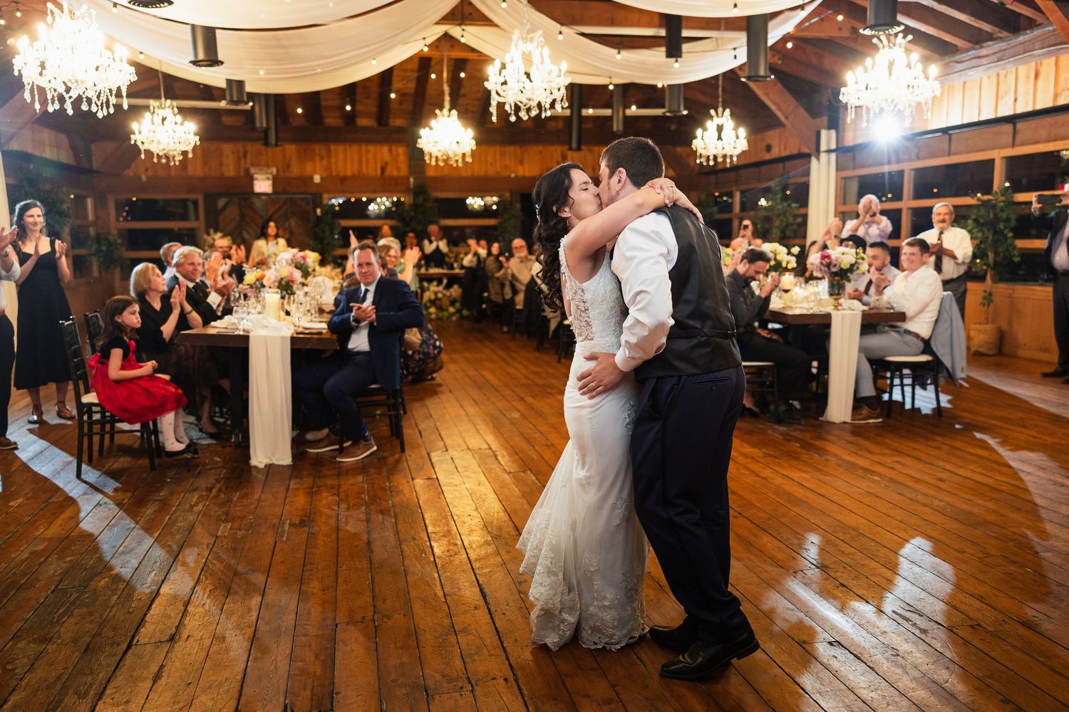 Bride and groom share first dance on wooden floor in rustic barn wedding reception with chandeliers and guests seated at tables.