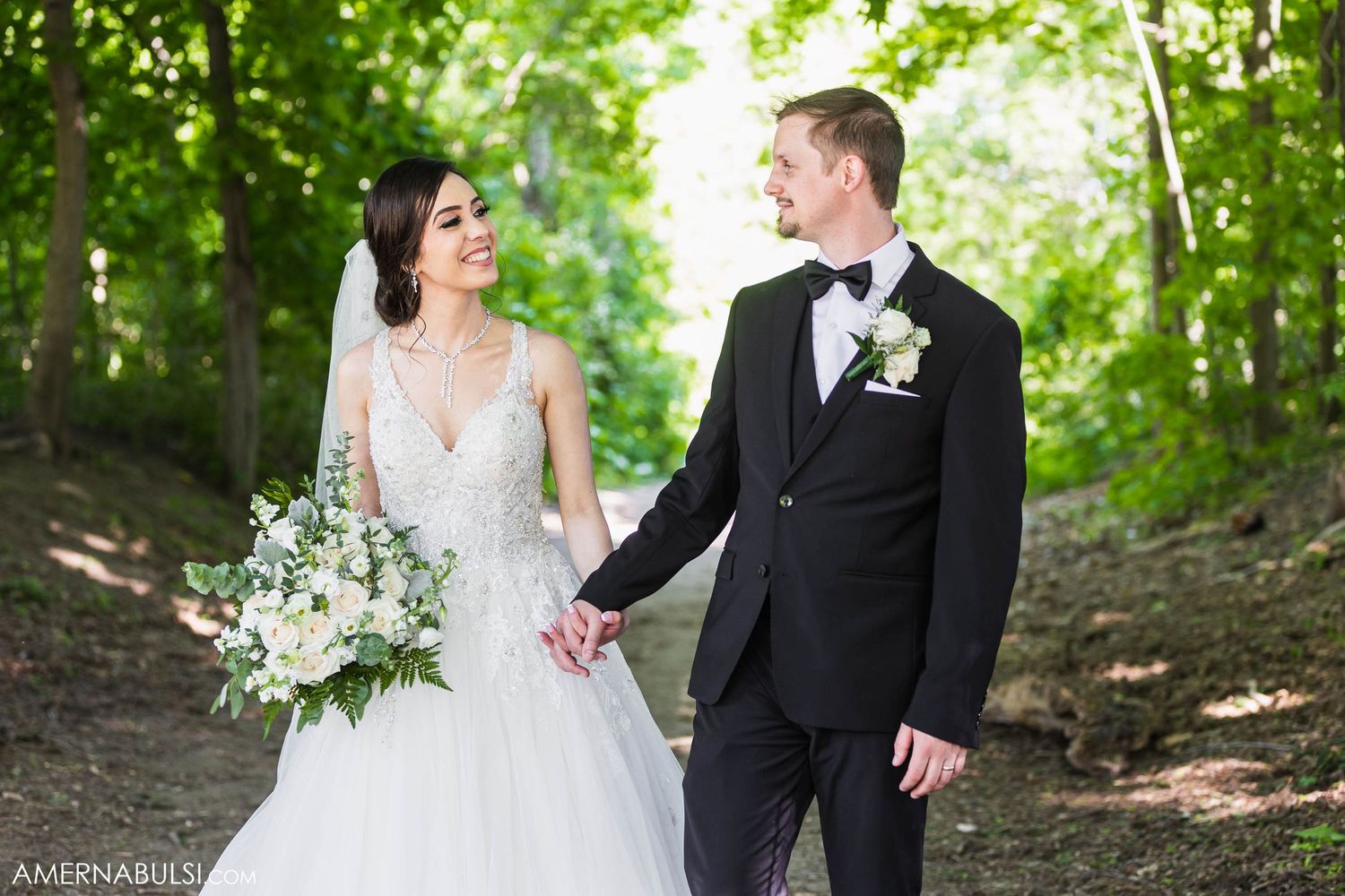 Couple shares loving glance while walking through sunlit forest path on wedding day.