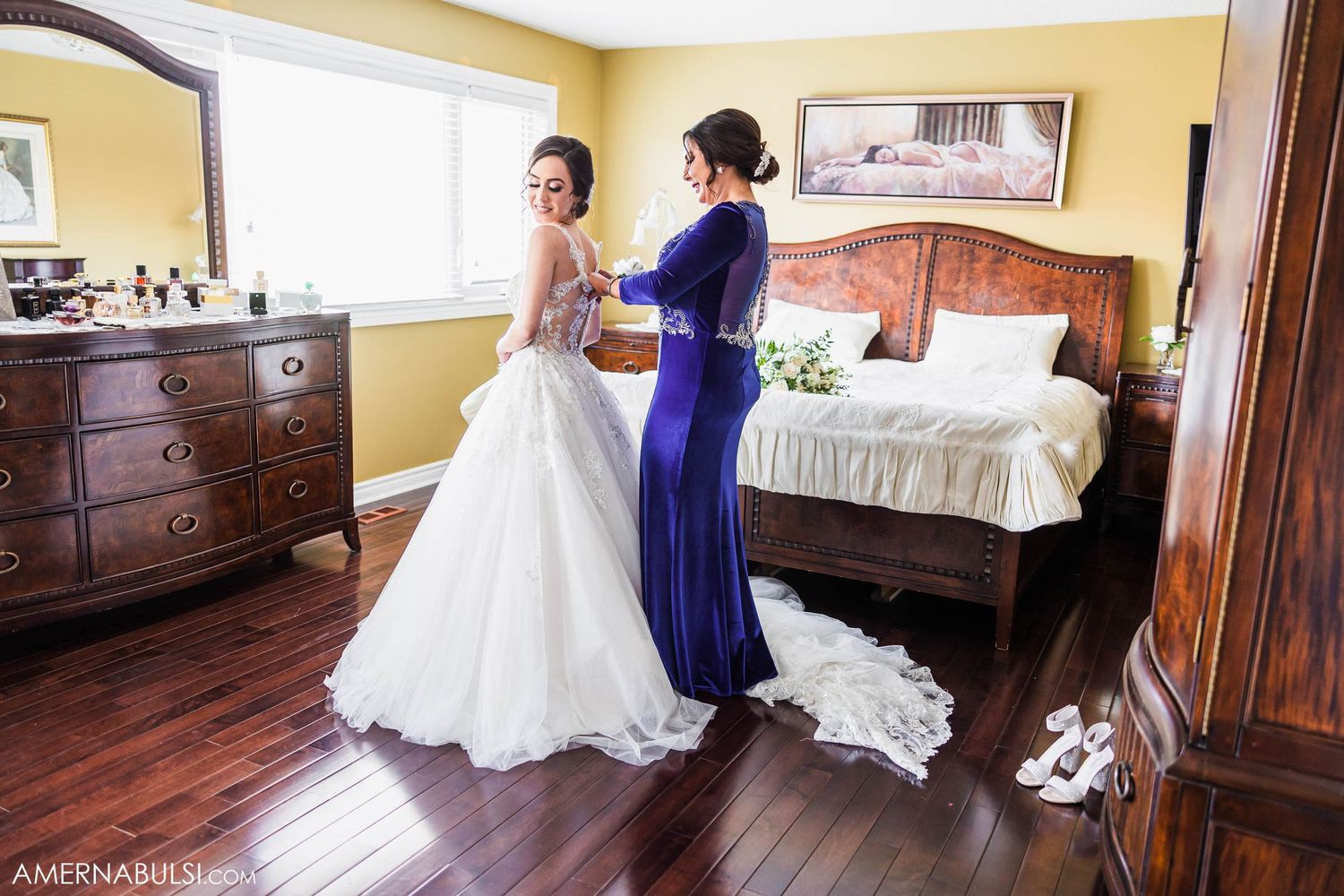 A bride in a white lace gown is helped with final adjustments in a bedroom with dark wood floors.