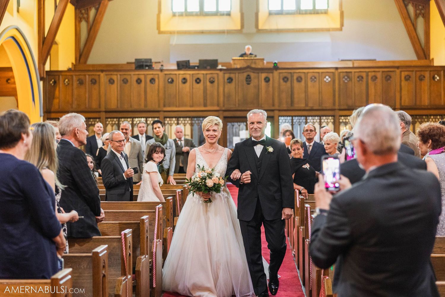 Wedding processional in a traditional church with wooden pews and red carpet.