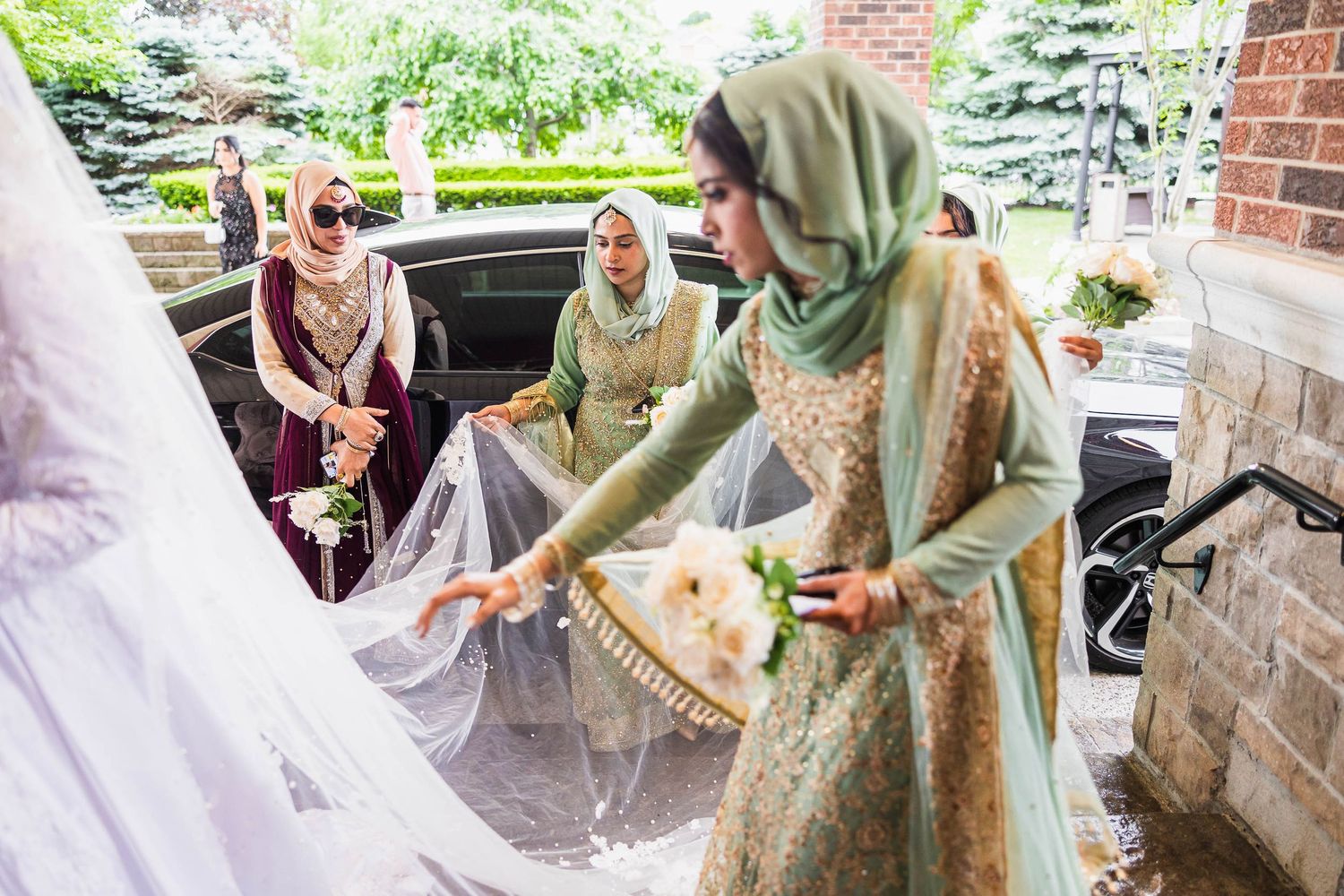 Women in traditional dress help arrange a wedding gown outside a brick building.