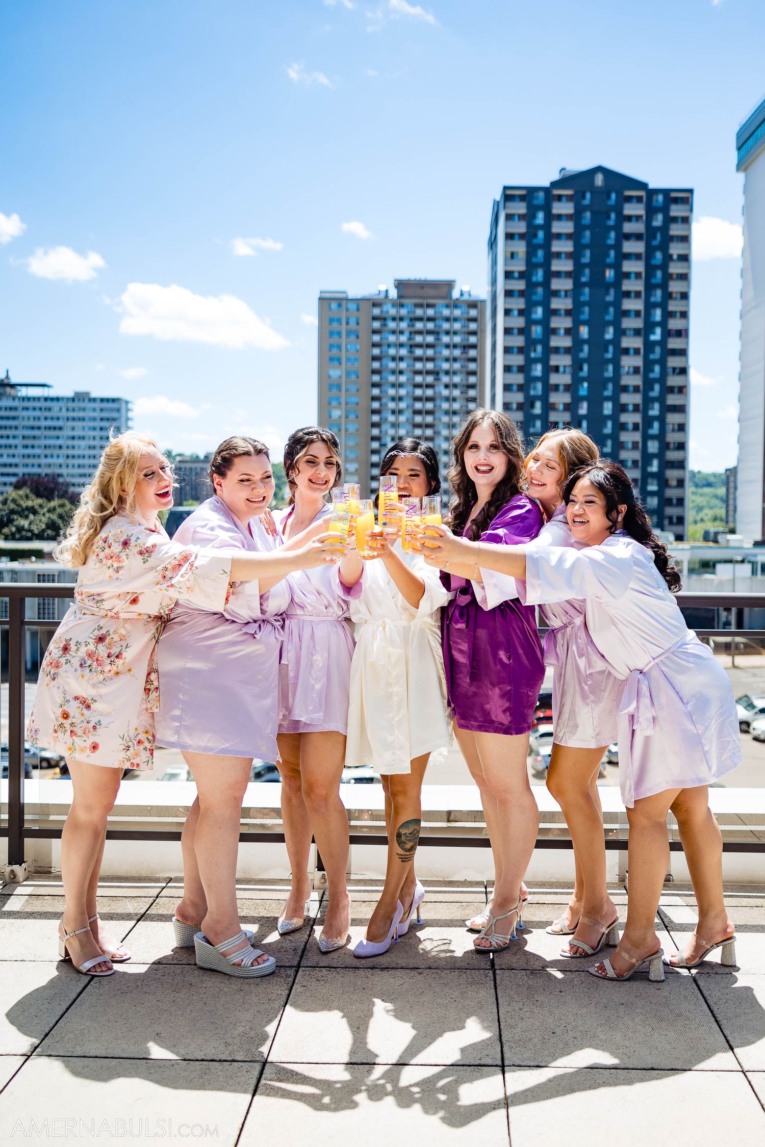 Wedding party in pastel robes clink glasses during rooftop celebration.