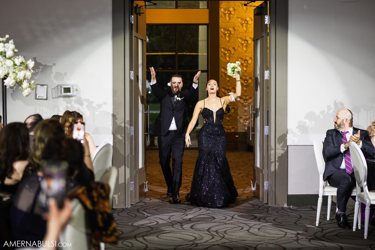 Wedding guests celebrate with raised arms as they enter through golden doorways at a reception.