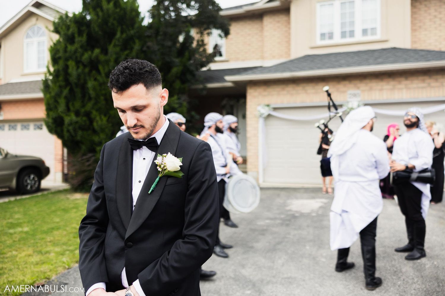 Groom in black tuxedo and bowtie stands outside suburban home while wedding party gathers in background.