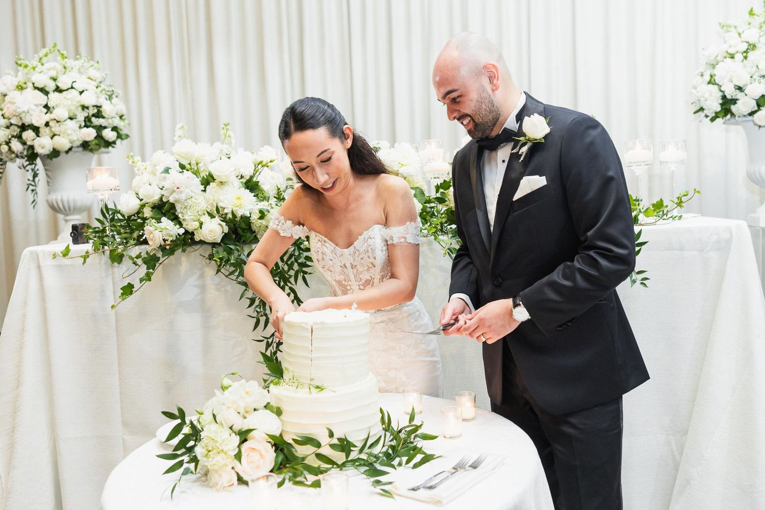 Newlyweds cutting their elegant white wedding cake adorned with greenery and florals at their reception.