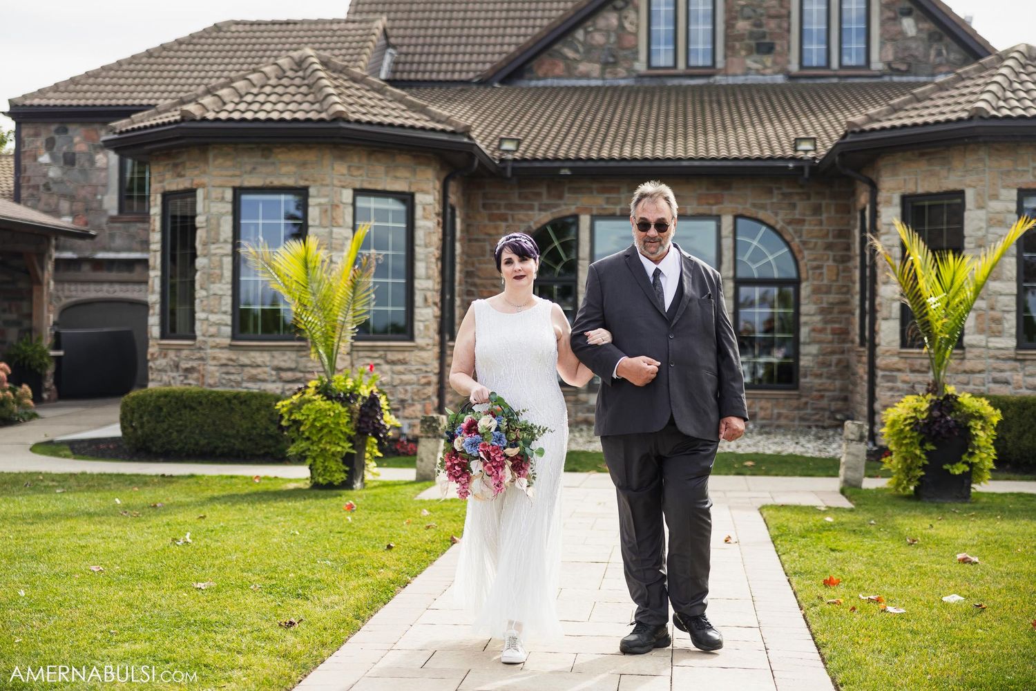 Couple walking down path in front of elegant stone house with arched windows.