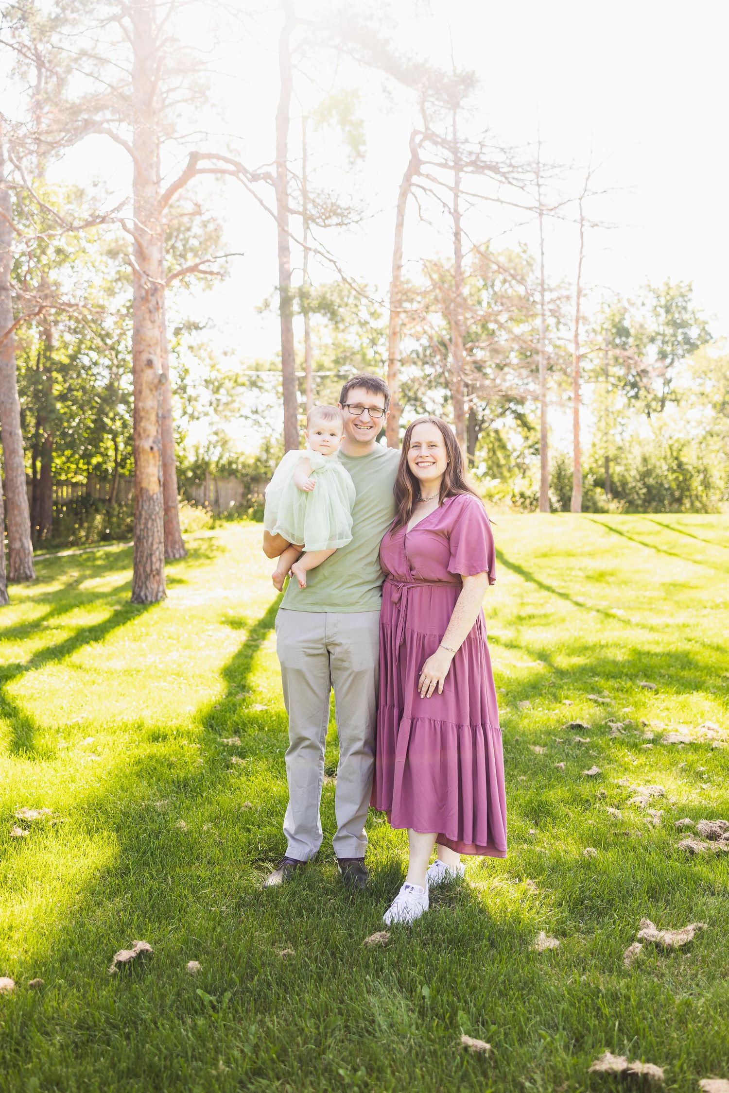 Couple in green shirt and mauve dress pose together in sunny pine forest.