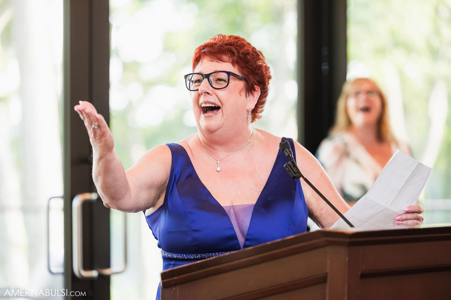 A guest in a blue dress gives an animated speech at the wedding reception podium.