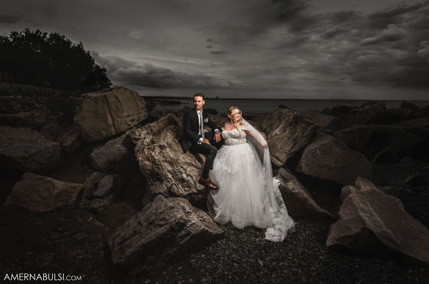 Dramatic wedding photos captured on dark rocky shoreline at dusk.