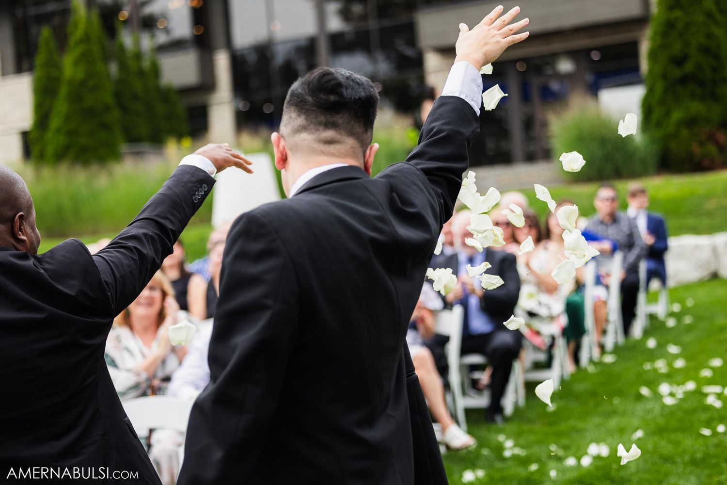 Wedding officiants raise their arms in celebration during outdoor ceremony with guests seated on lawn.