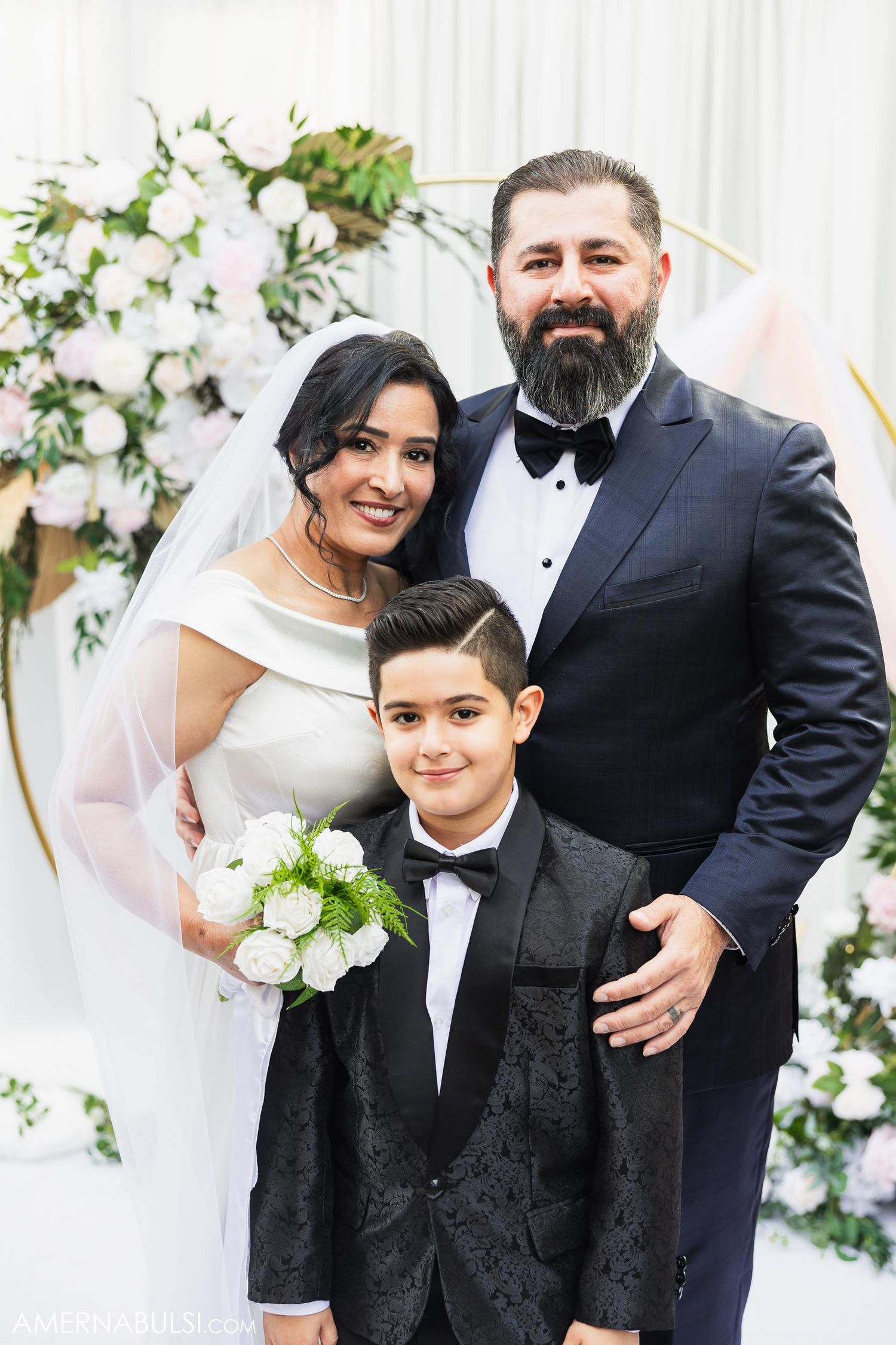 A wedding portrait of newlyweds and young boy in formal attire against floral backdrop with white roses.