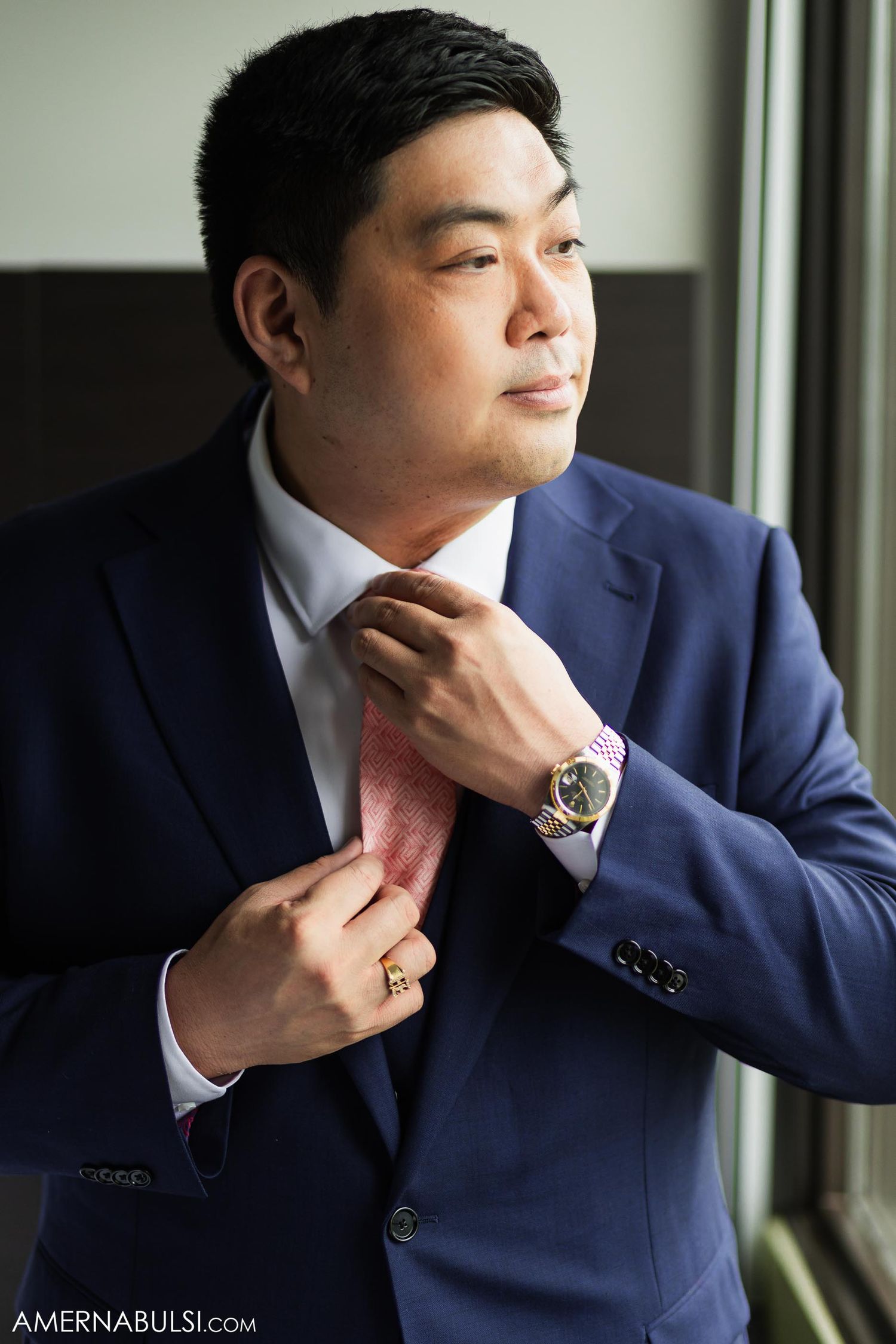 Professional wearing a navy suit and pink tie adjusts his collar while looking towards a window.