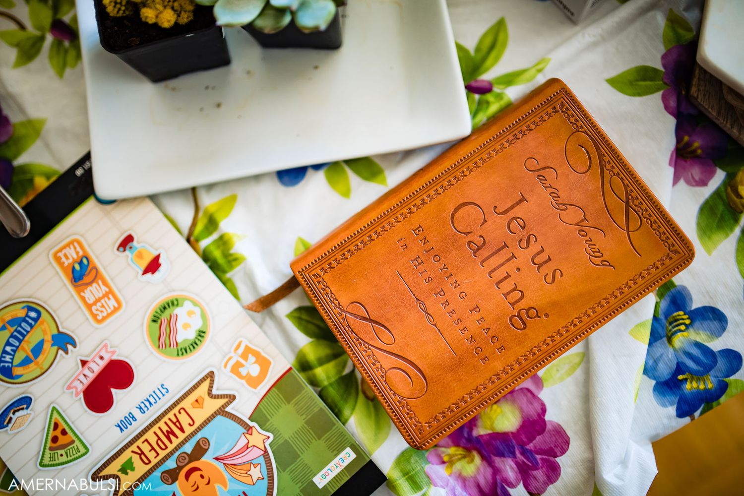 Leather wallet with embossed designs photographed among colorful flowers and greenery on white surface.