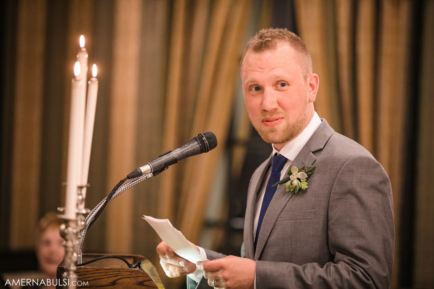 Groomsman in grey suit delivering a wedding speech at reception podium.