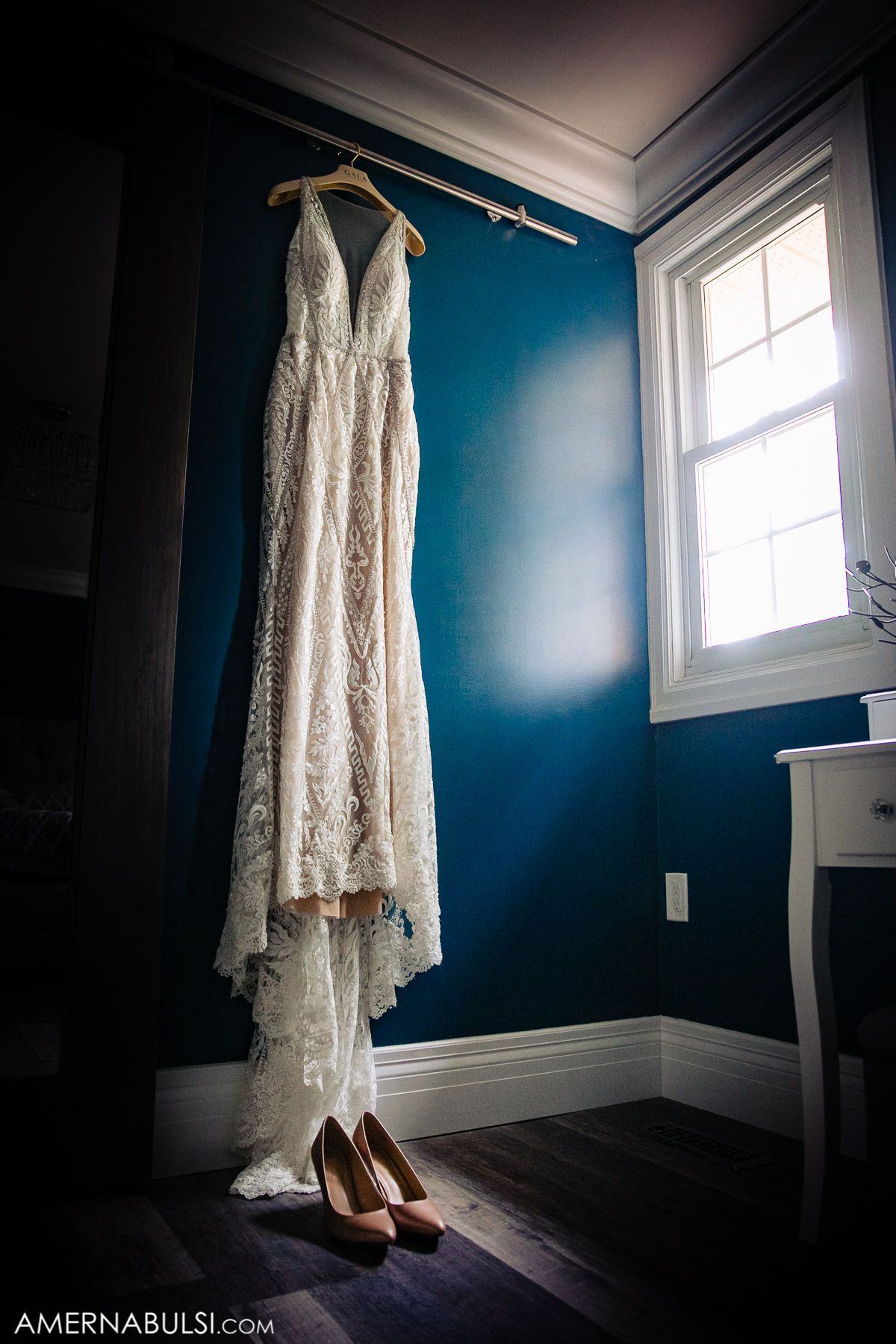 White lace wedding dress and nude heels displayed against deep blue walls in a sunlit room.