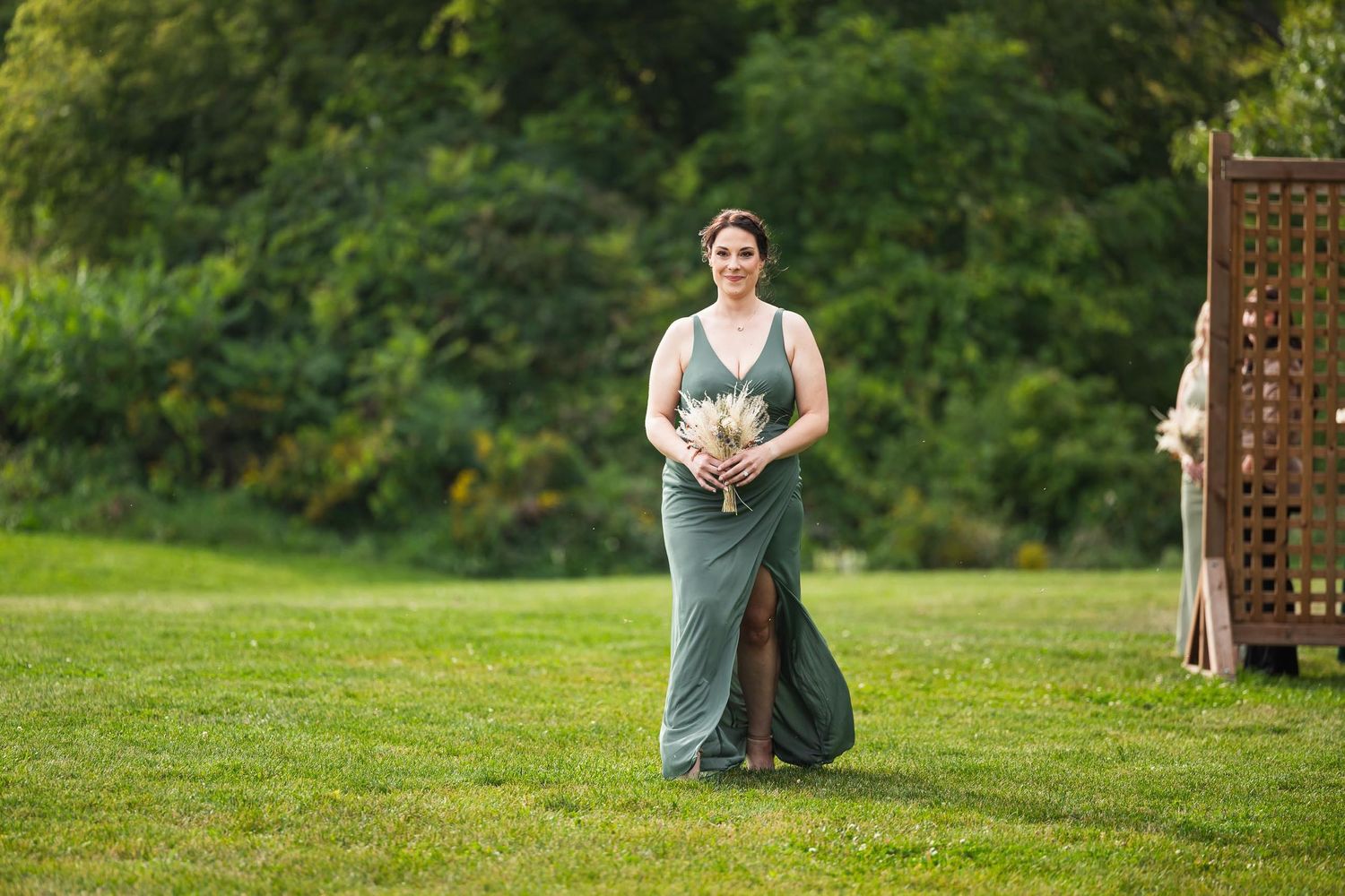 Bridesmaid in sage green dress walks down grassy aisle holding bouquet.