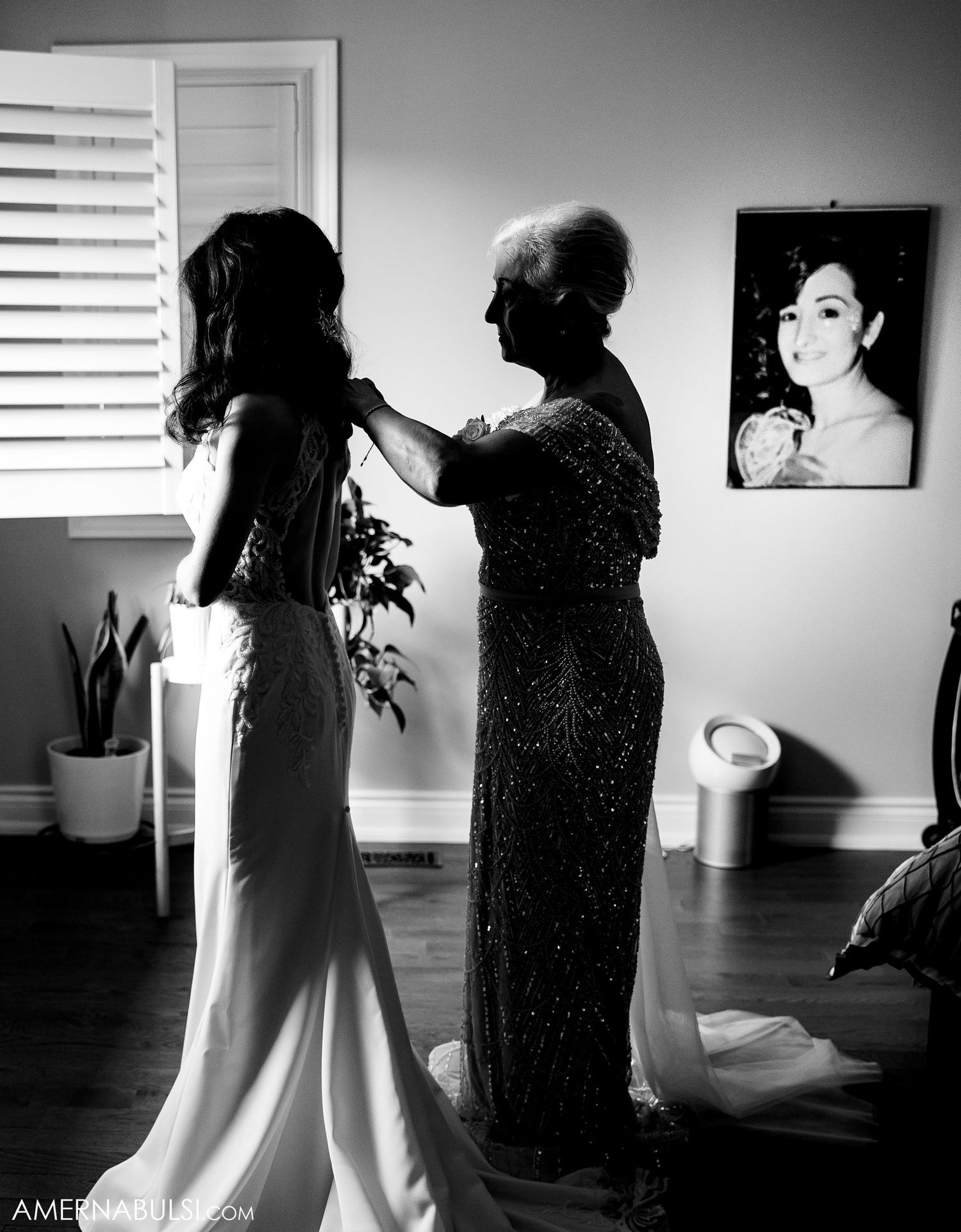 A black and white photo showing someone helping another person with a wedding dress in a room with a window.
