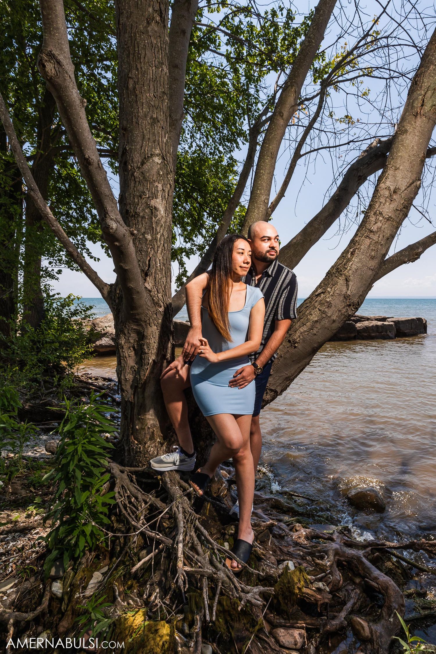 A couple poses together on a tree trunk by the rocky shoreline at sunset.
