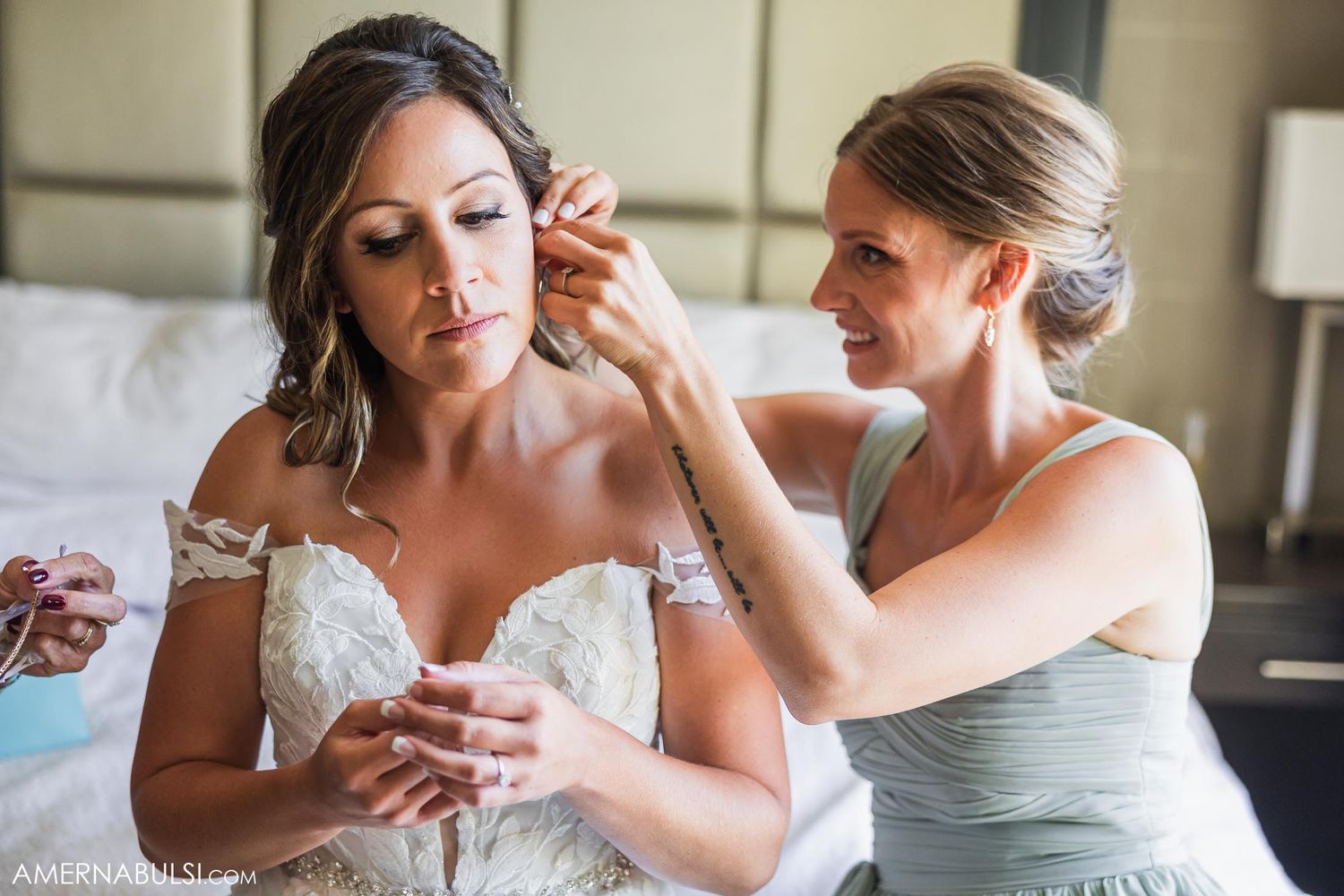 A bride getting help with earrings during wedding preparations in a soft lit room.