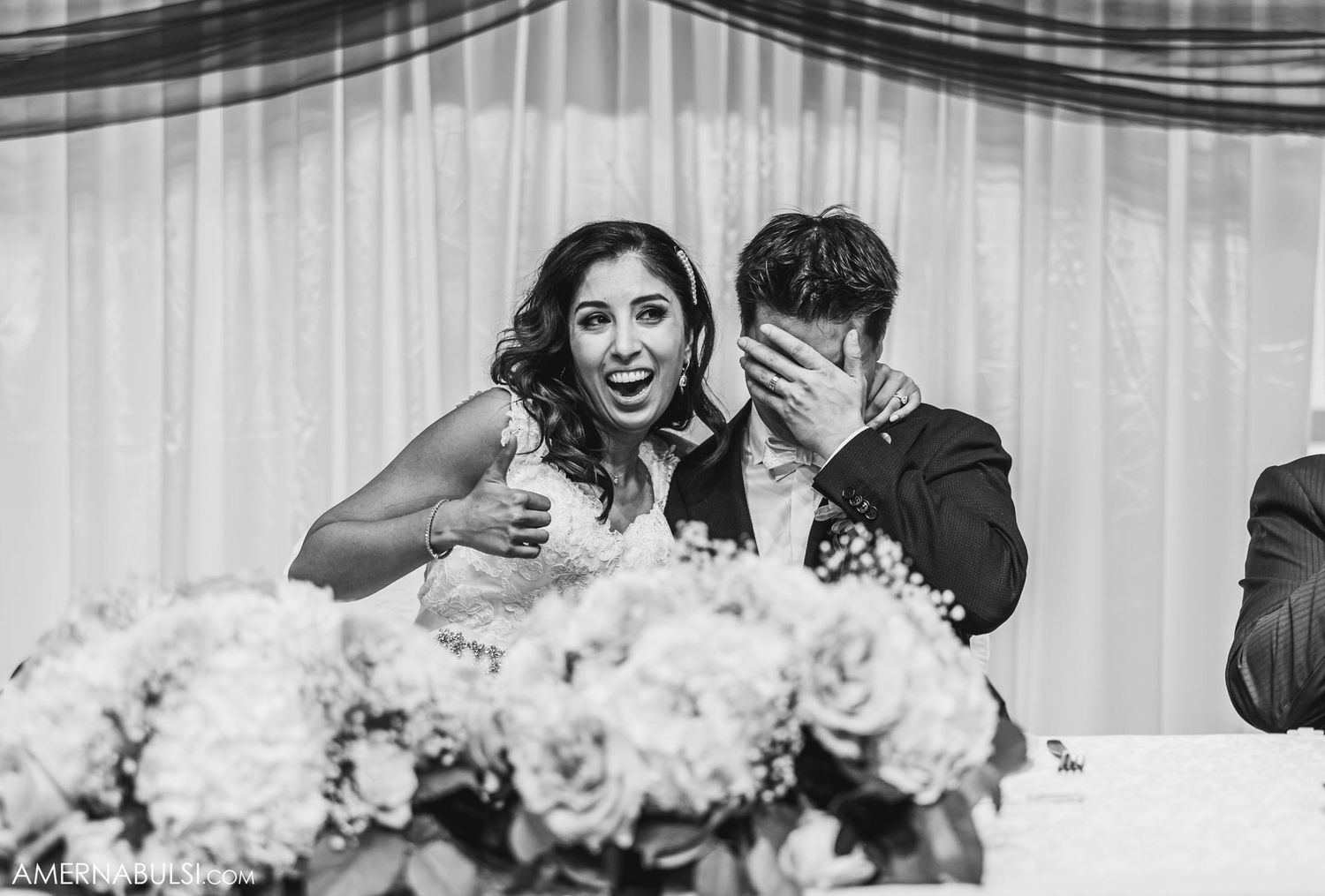 Candid black and white photo of newlyweds sharing a joyful moment at their reception head table.