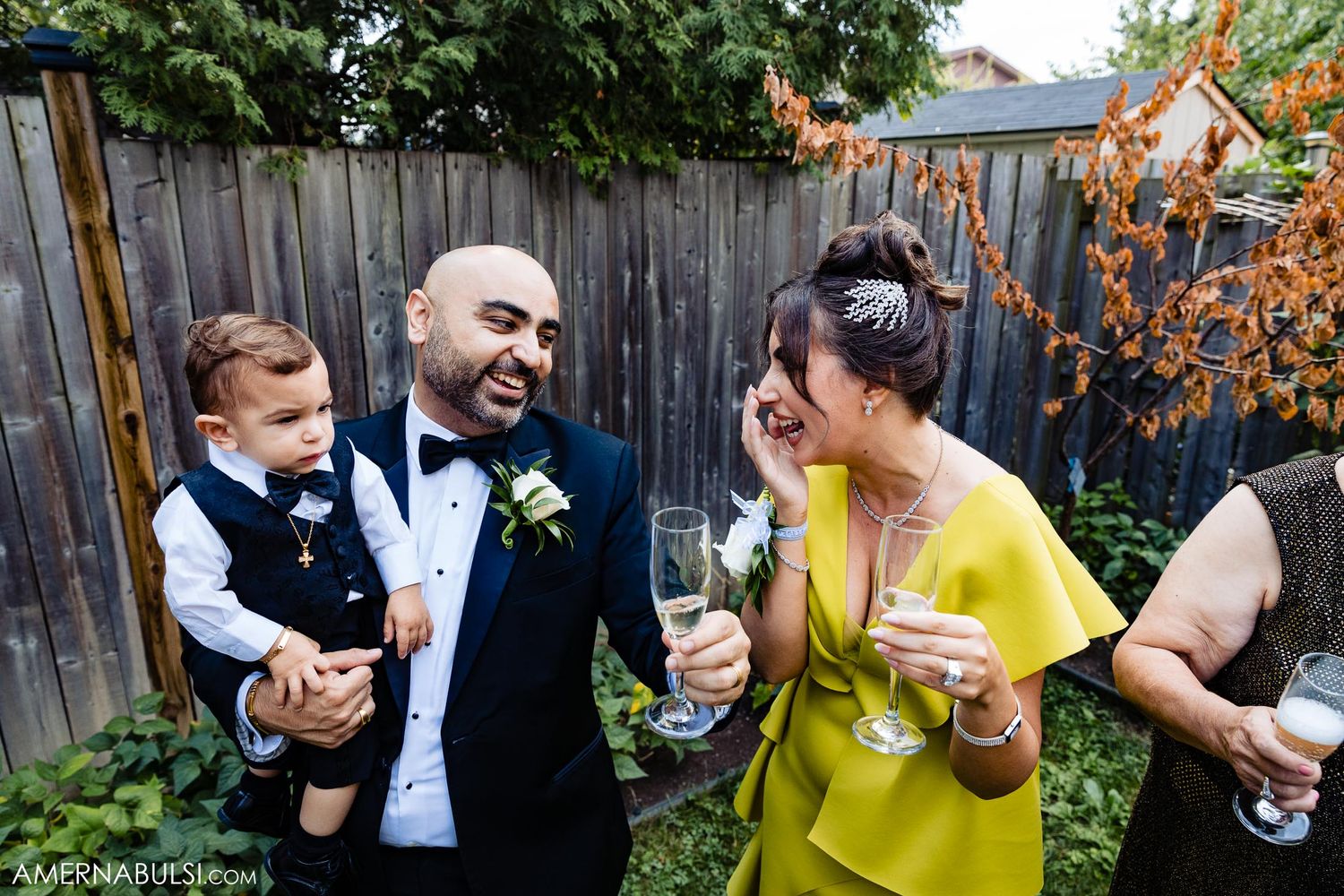 Wedding guests share laughter and drinks in a backyard garden setting with rustic wooden fence.