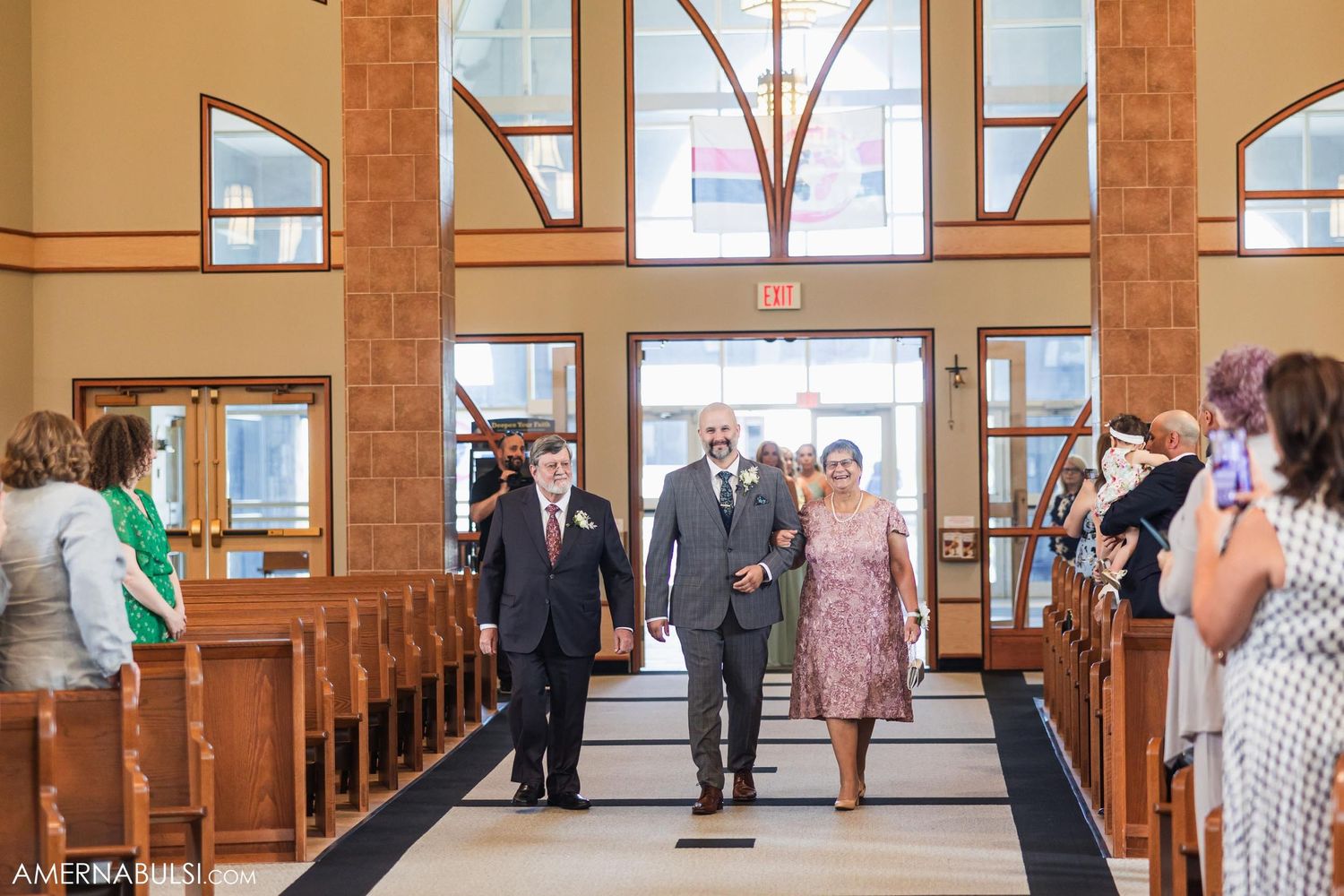 Wedding party members walk down a church aisle while guests watch from wooden pews.
