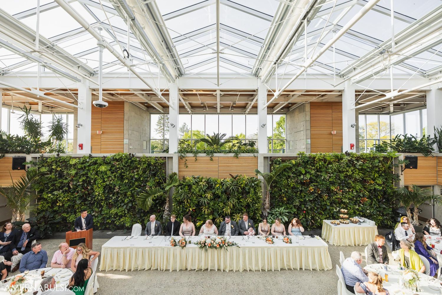 An outdoor wedding reception setup under a glass conservatory roof with green wall decorations.