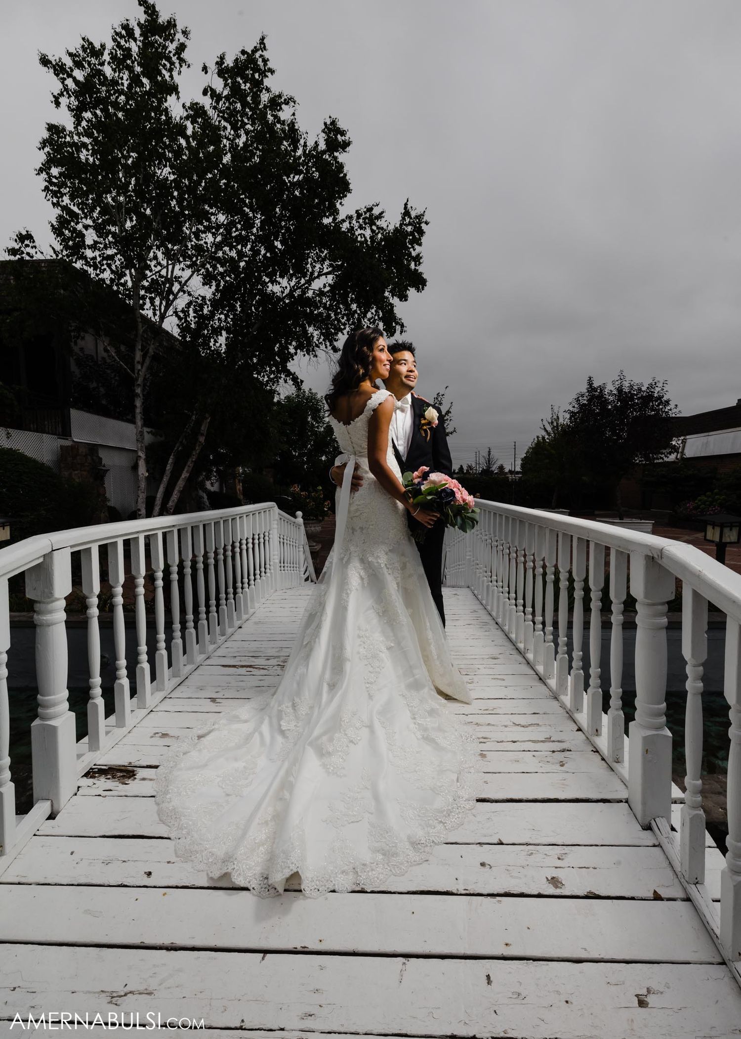 Newlyweds pose dramatically on a white bridge walkway with the bride's long lace dress trailing behind.