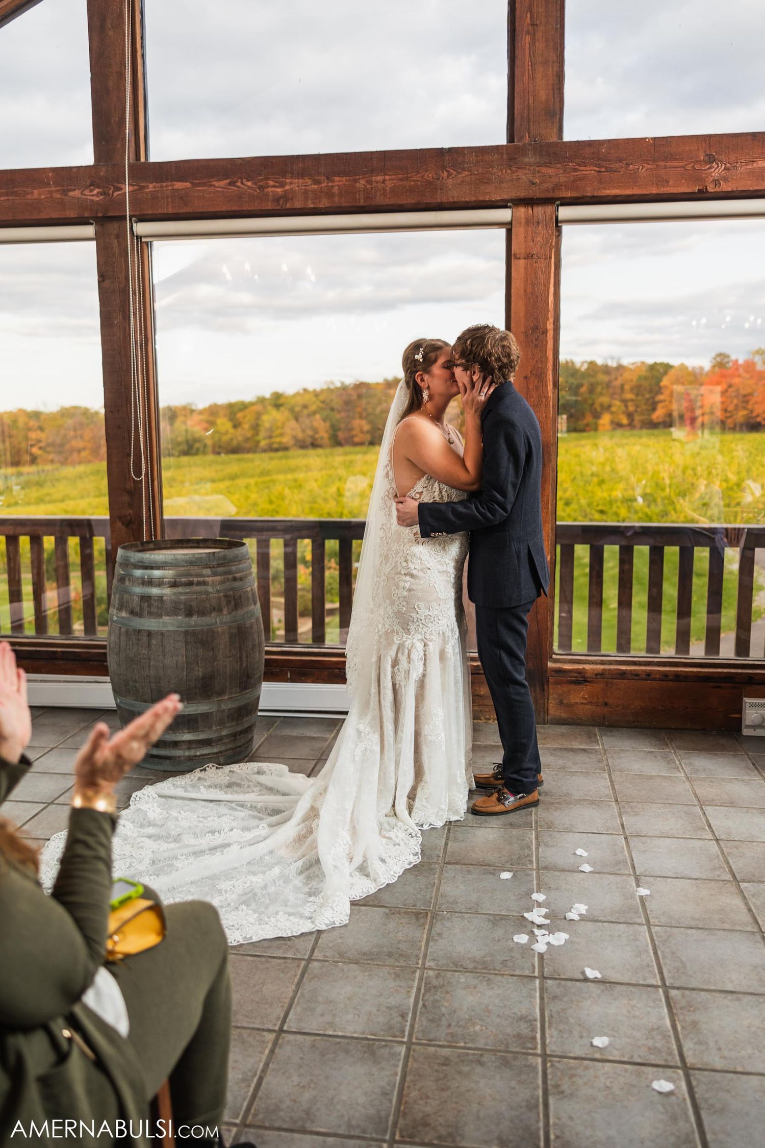 Wedding couple shares first kiss in rustic venue with wine barrel and autumn views.