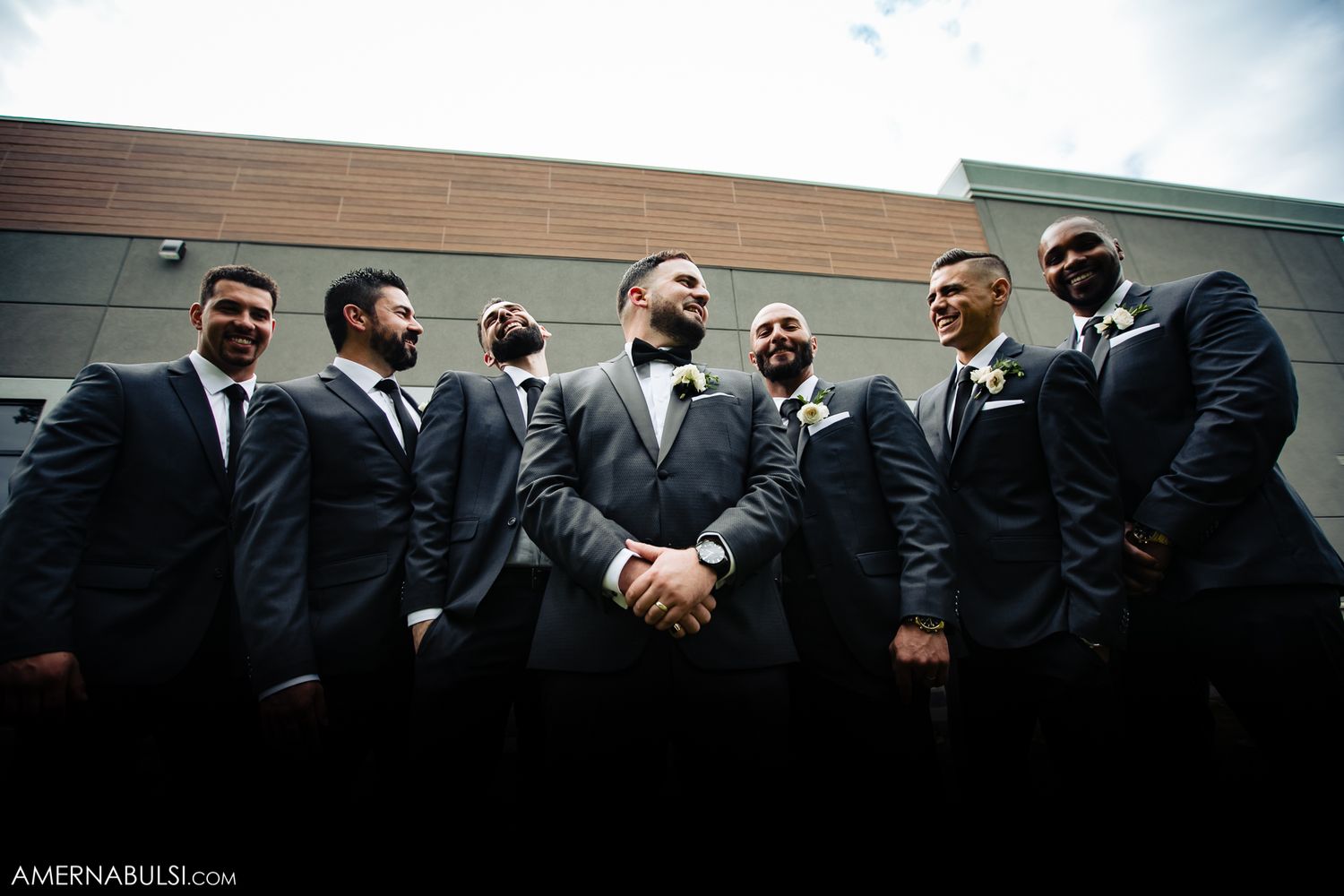 Groomsmen in black suits posing together against building exterior.