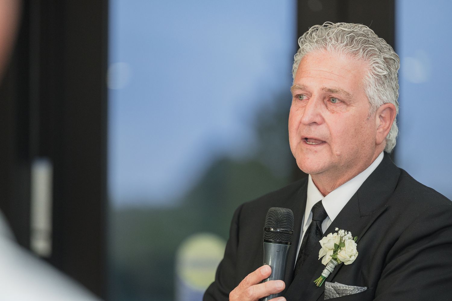 Older man in black suit with white boutonniere speaking into microphone at formal event.