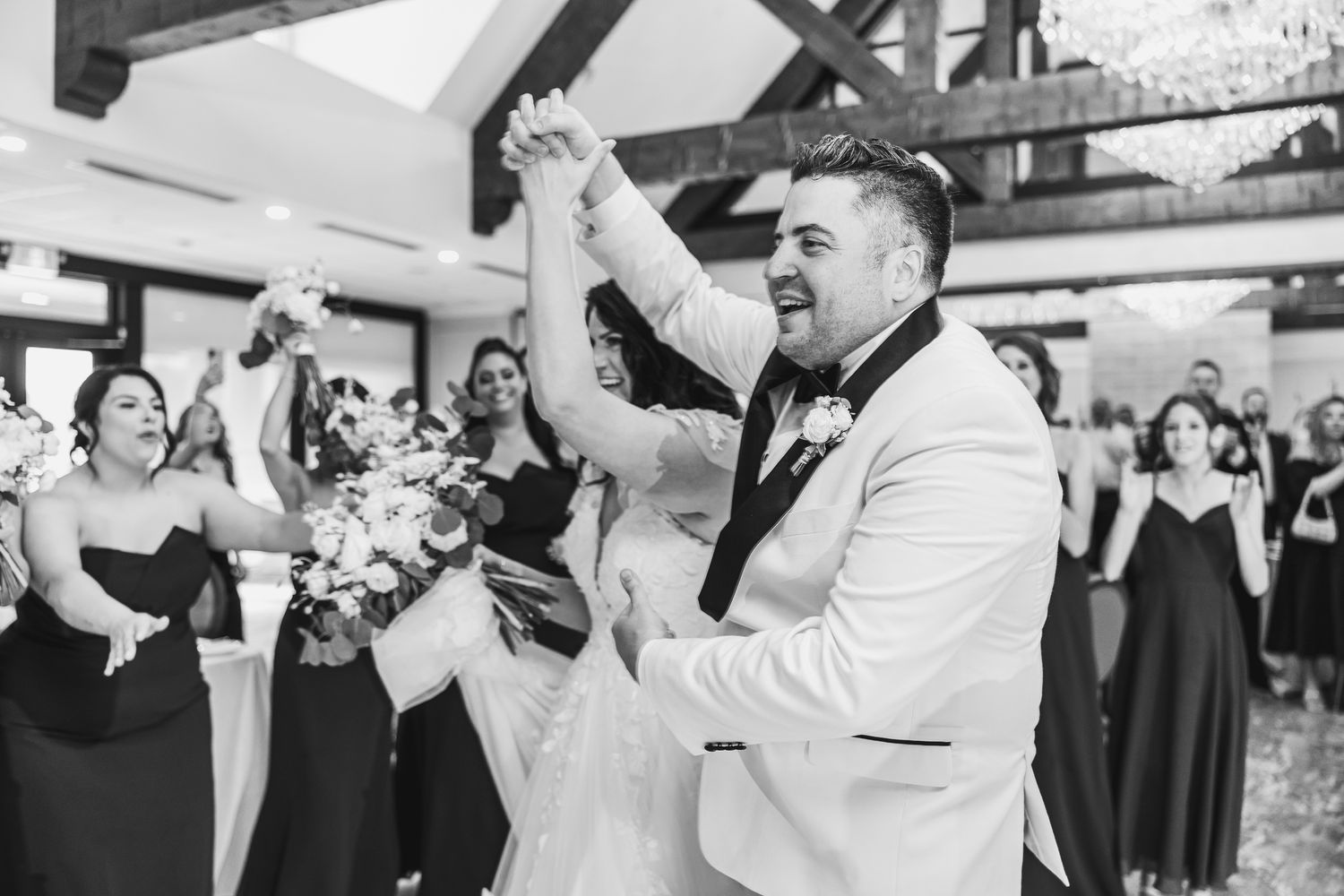 Joyful groom in white tuxedo celebrates with bride and bridesmaids in black dresses at wedding reception.
