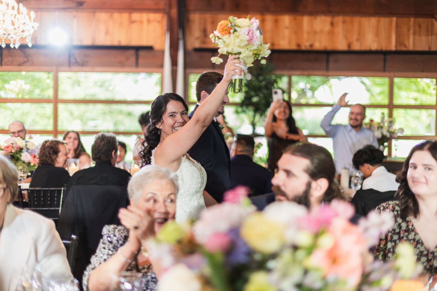 Bride raises her colorful bouquet triumphantly while smiling guests celebrate around her at the reception.