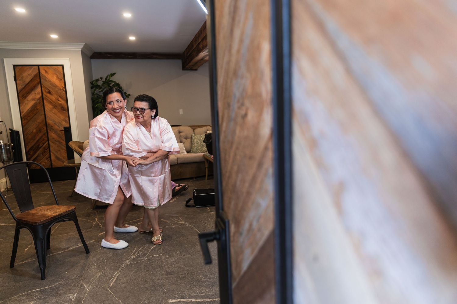 Two women in pink robes laughing and getting ready together in a modern bridal suite.