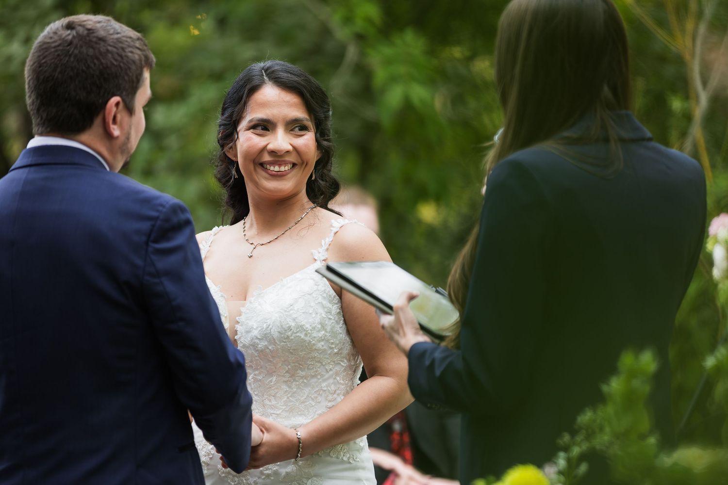 Smiling bride holds hands with groom during outdoor wedding ceremony officiated by female officiant.
