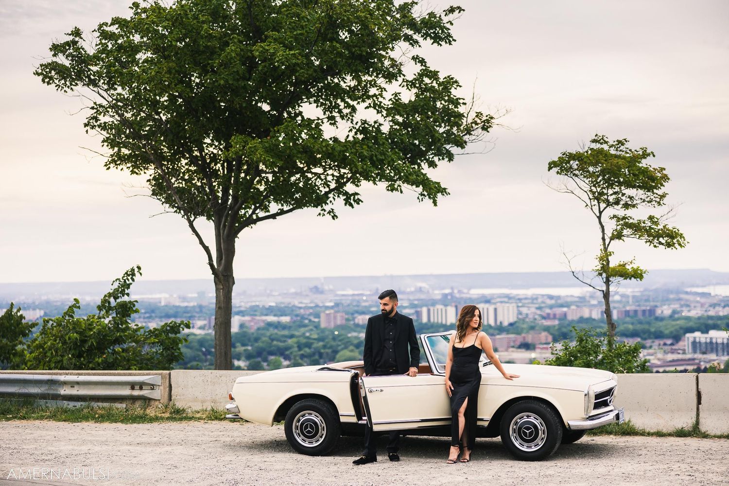 Scenic wedding photo series with vintage car under trees overlooking city vista.