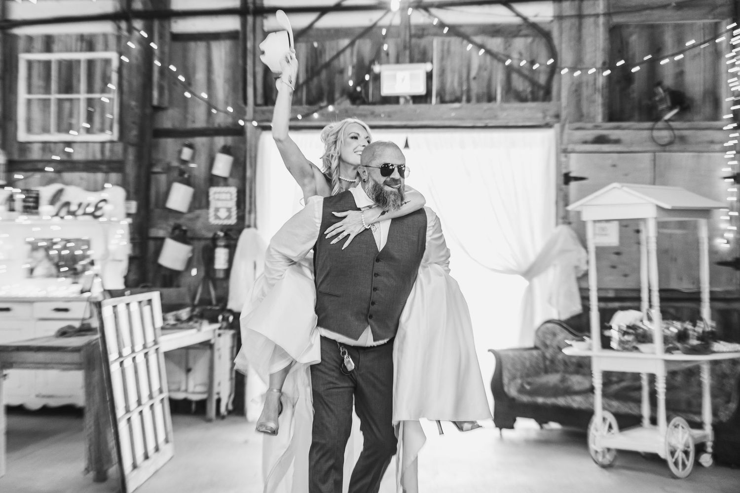 Black and white photo of a joyful couple dancing at a rustic wedding venue with string lights.