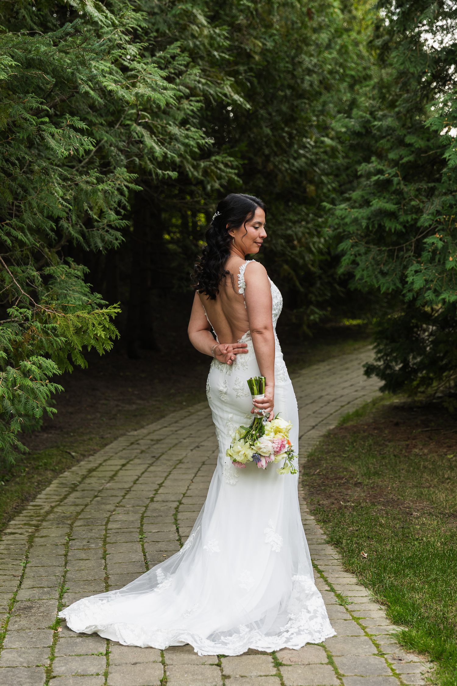 Bride in elegant backless lace wedding gown holding bouquet while walking on a brick garden path.