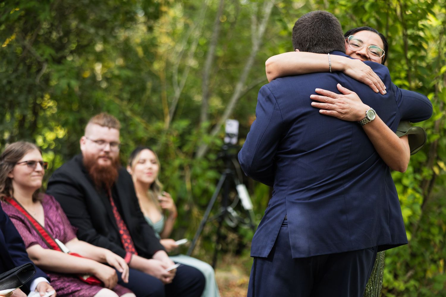 Two men share an emotional hug at an outdoor wedding ceremony while guests watch in the background.