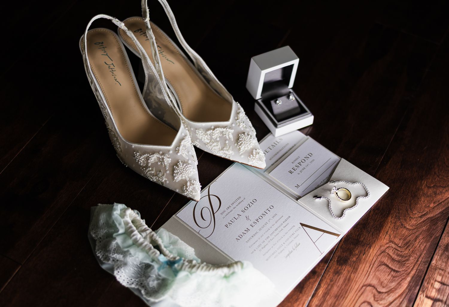 Elegant wedding flat lay featuring lace bridal heels, ring box, garter, and wedding invitation on dark wood.
