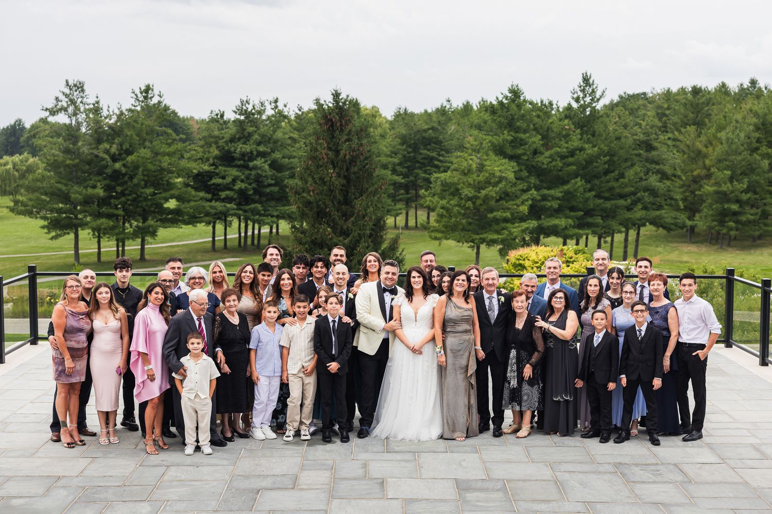 Large wedding family group photo with bride and groom surrounded by guests on stone terrace.