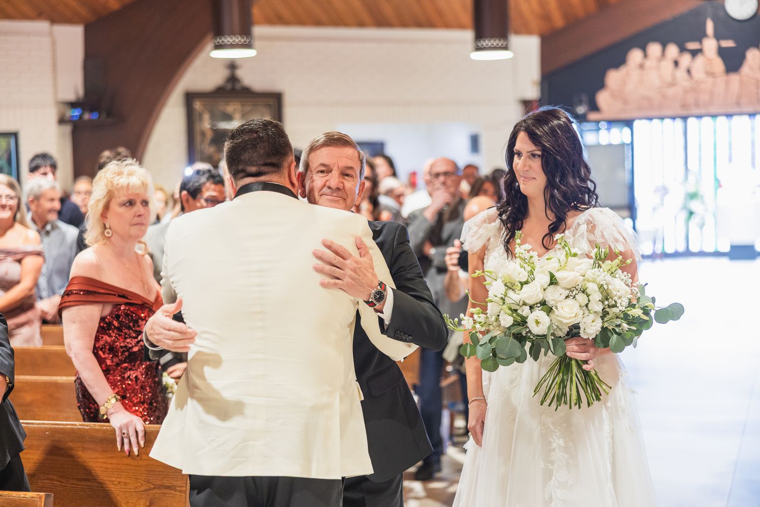 Groom embraces a guest as bride holding white bouquet watches during church wedding ceremony.