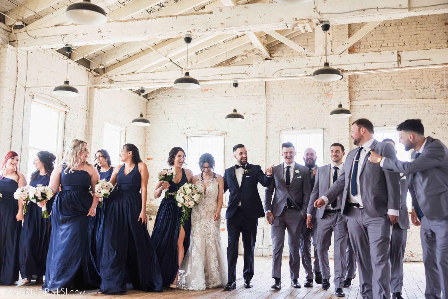 Wedding party laughs together in bright industrial space with exposed brick and beams.