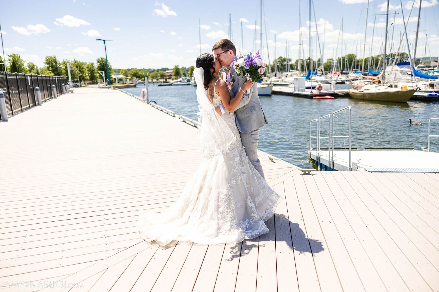 Newlyweds embrace on a sunny marina boardwalk surrounded by docked sailboats.