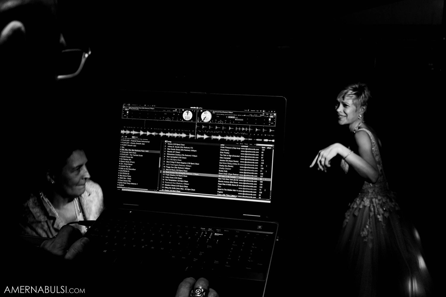 A black and white photo showing a DJ setup with a computer screen displaying playlists in a dark environment.