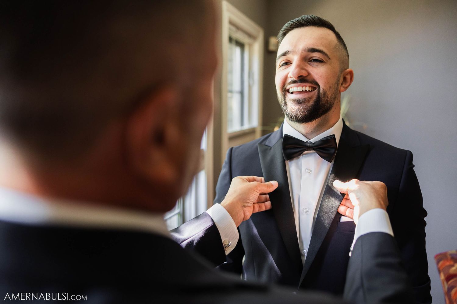 A groom smiles joyfully while someone adjusts his bowtie during wedding preparations.