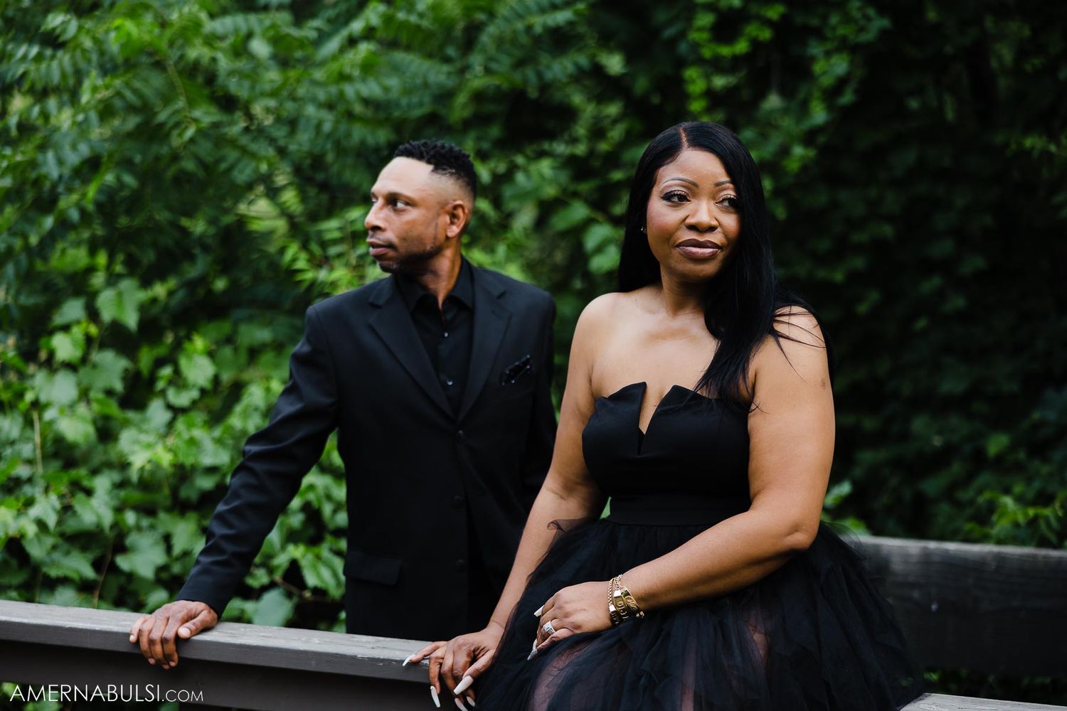 Stylish couple in black attire pose together on a bridge surrounded by lush greenery.