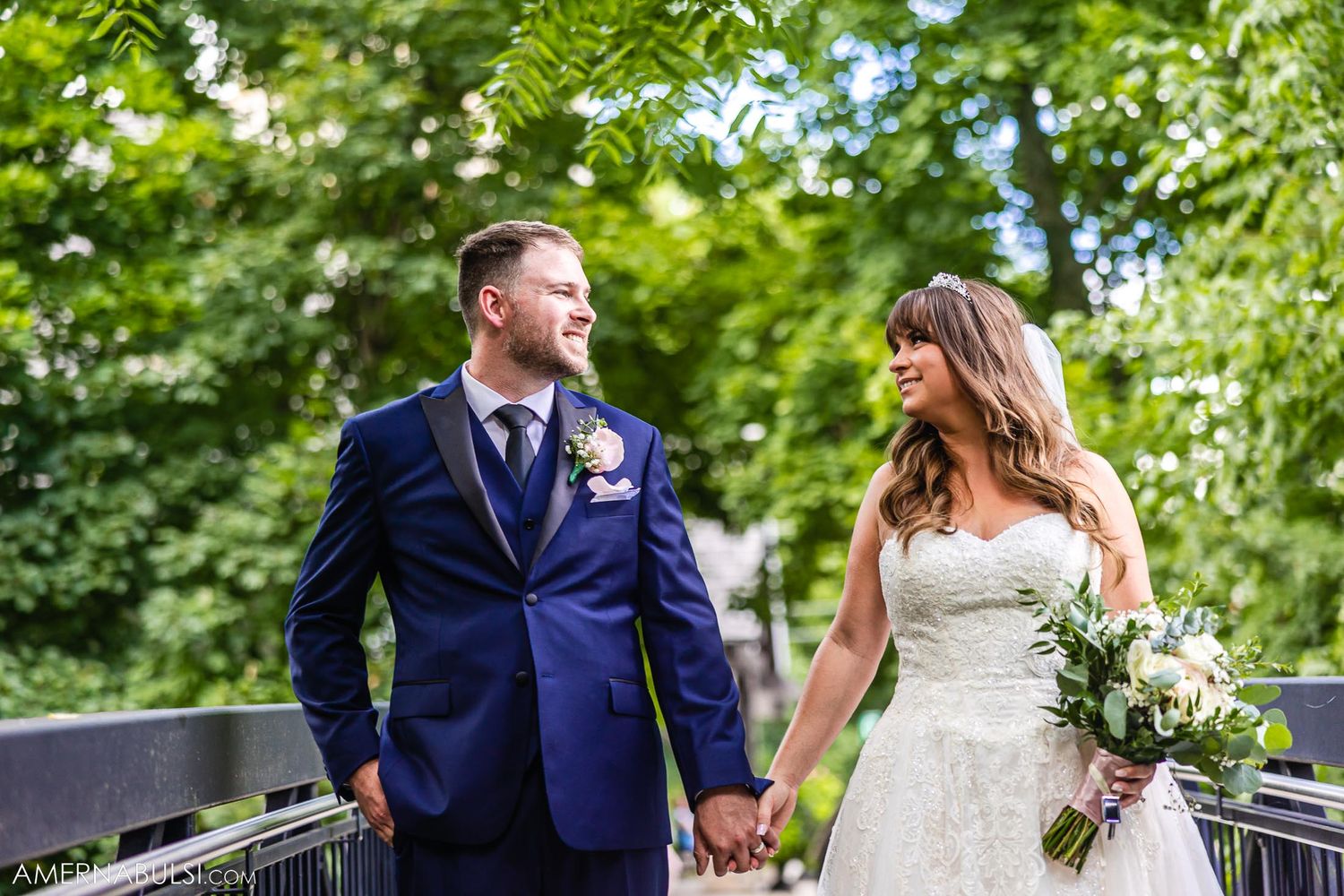 Newlyweds hold hands while standing on bridge surrounded by summer greenery.