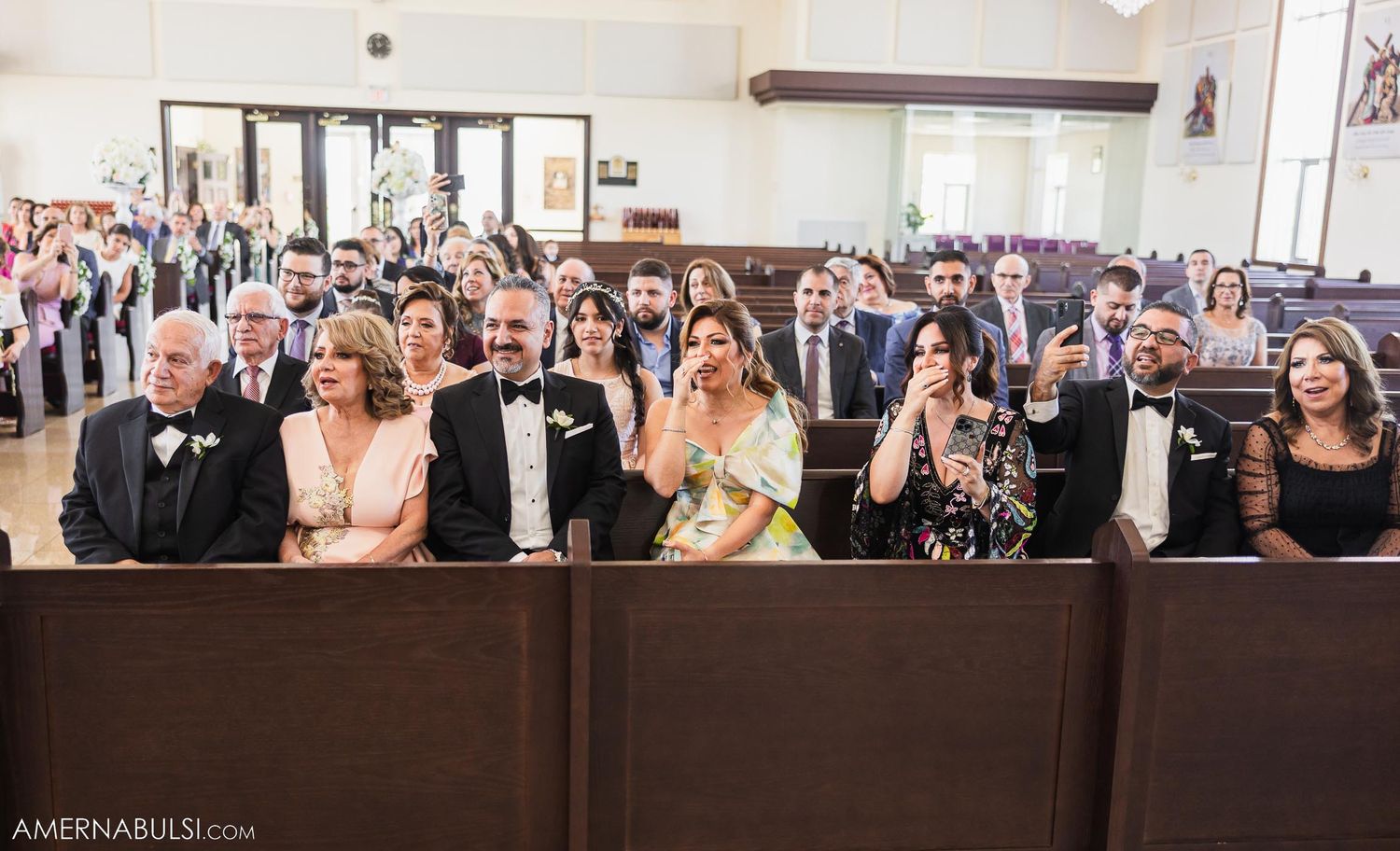 Wedding guests in formal attire seated in church pews watching ceremony with joyful expressions.